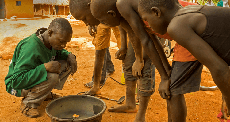 Miners burn mercury off the gold amalgam in the open air close to their dwellings to produce a piece of gold which is sitting on top of the match box. The gold is being inspected by the middle man (left) and mine workers.