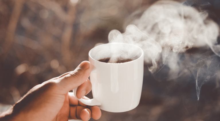 Hand holding a mug of coffee with steam billowing out and a winter tree background