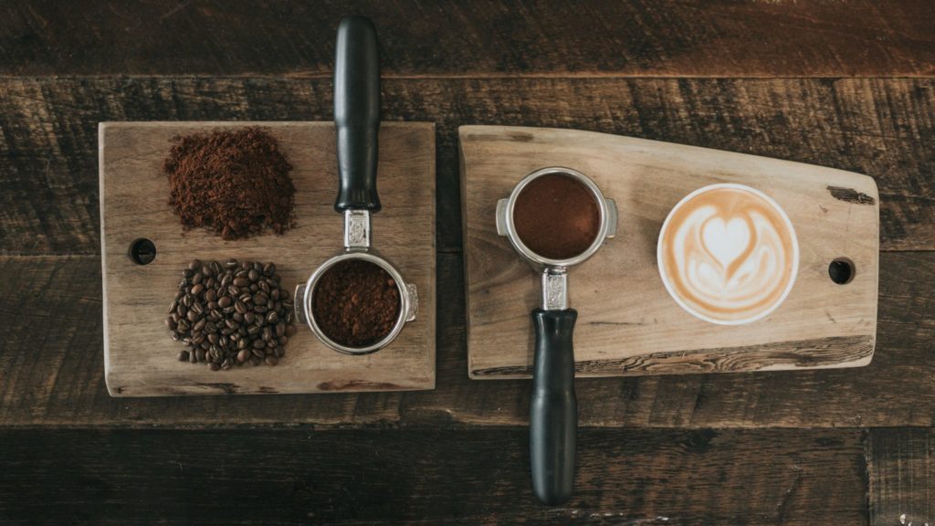 Shows ground coffe, coffee beans and a cup of coffee on a wooden board, shot from above
