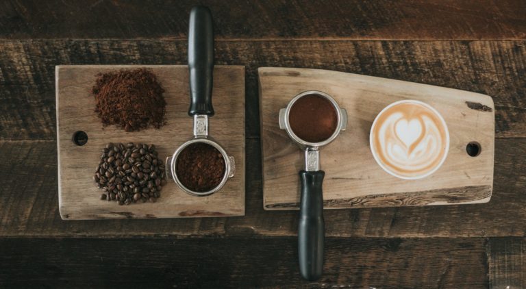 Shows ground coffe, coffee beans and a cup of coffee on a wooden board, shot from above
