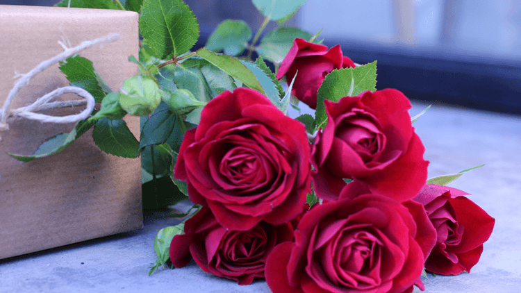 Red roses lying next to a present wrapped in brown paper with a string bow.