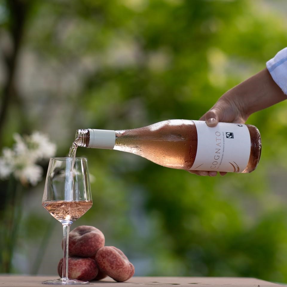Cognato rose wine bottle being poured into a wine glass. Pile of peaches on the table, green blurry nature in the background.