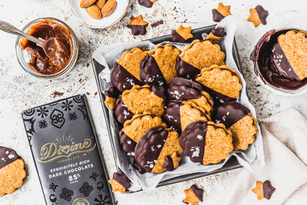 a plate of home backed sandwiched biscuits dipped in chocolate next to bar of divine chocolate