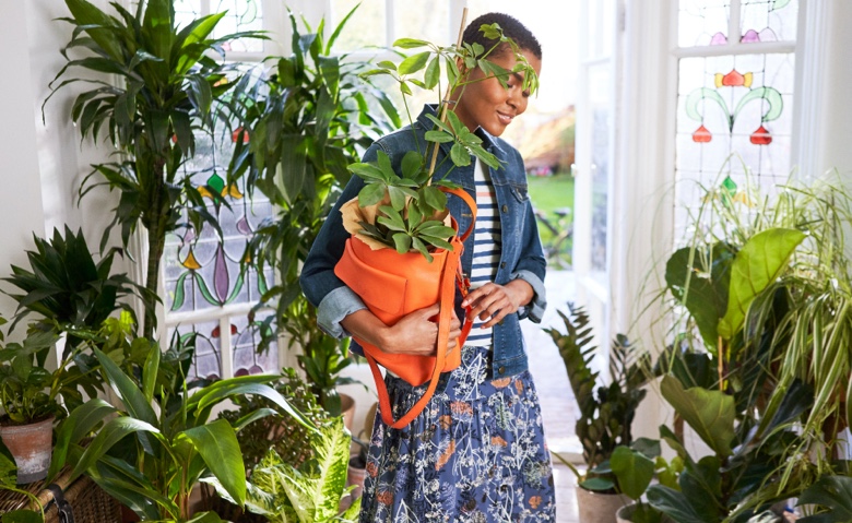 Woman in blue and pink striped t-shirt, denim jacket and flora design skirt holding a plant in an orange handbag, surrounded by lots of houseplants.