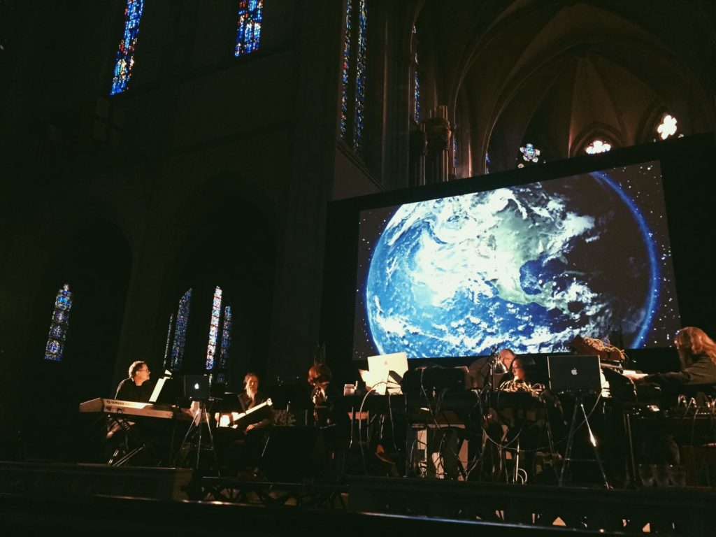 Orchestra on stage with a large screen behind them showing a satellite view of Earth