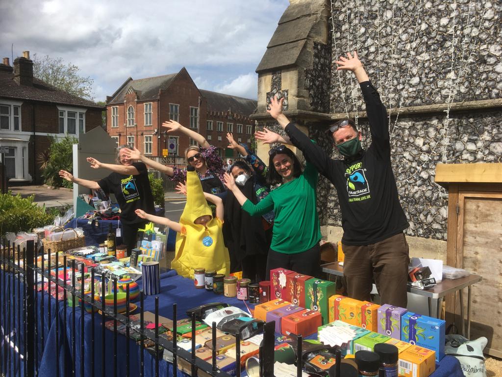 London Fairtrade campaigner group, waving their arms and hands to the right, standing in front of a stall selling Fairtrade products