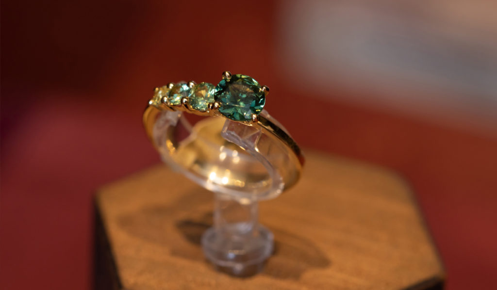 18ct Fairtrade yellow gold asymmetric green sapphire ring displayed on a wooden plinth with a red blurry table top background