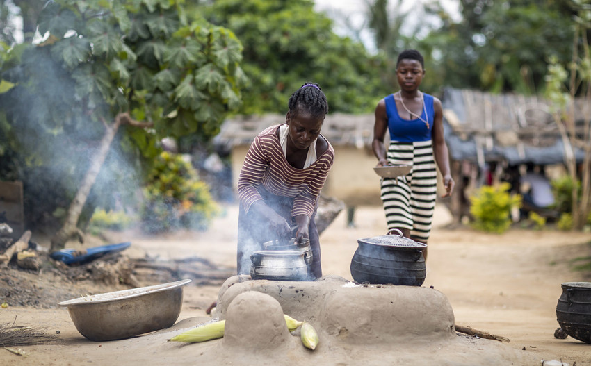 Therese preparing food in two pots, trees in the background and another woman approaches Therese 