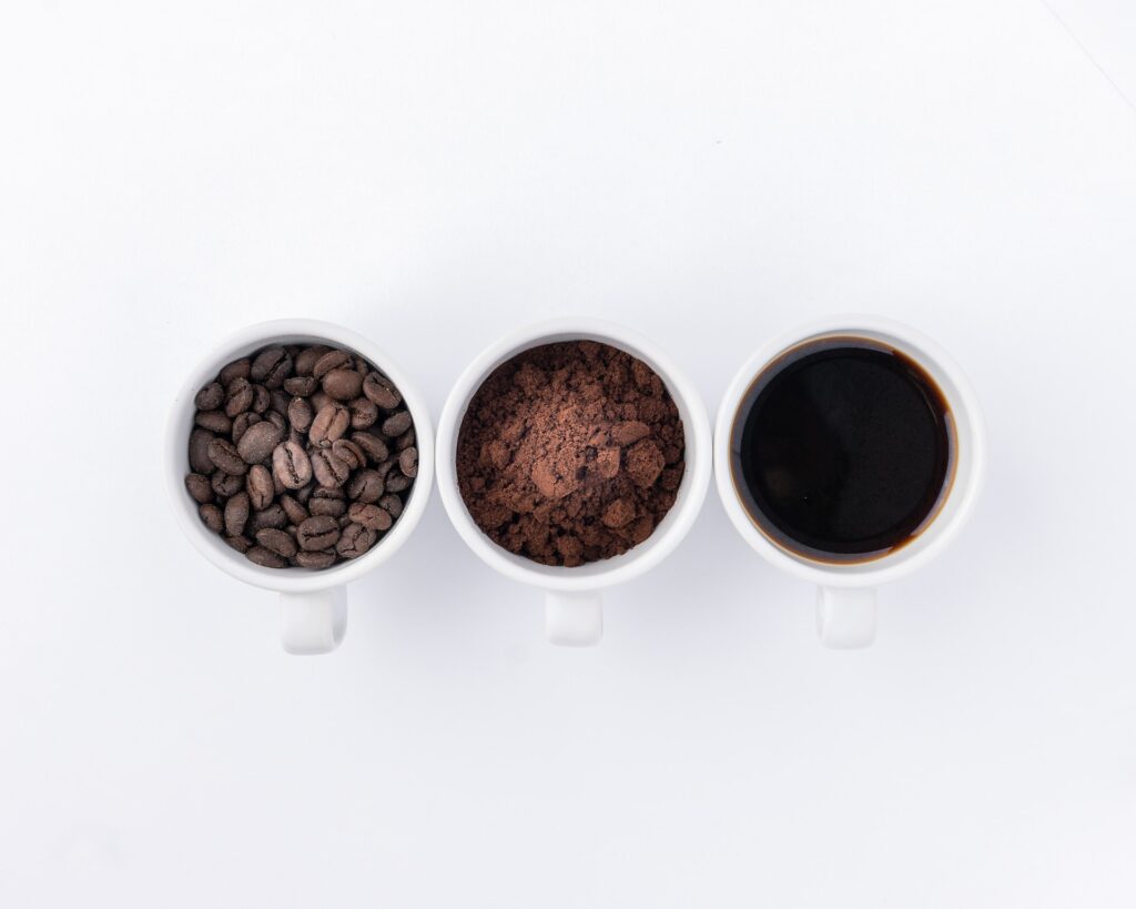 birds eye view of three white cups of coffee, first filled with coffee beans, second with powdered coffee granules, the third with black coffee drink