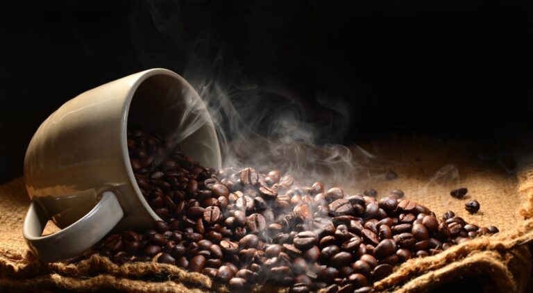 a cup on its side filled with coffee beans spilling out with steam to signify a hot cup of coffee