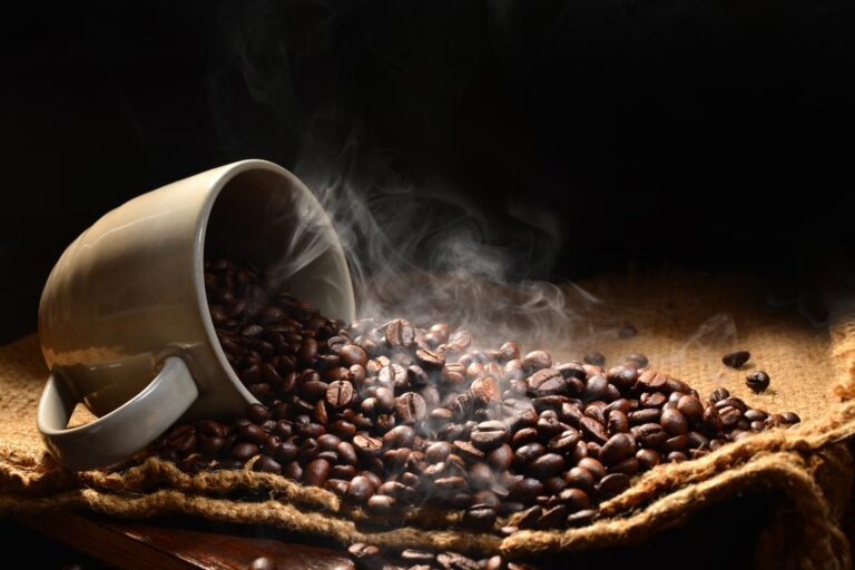 a cup on its side filled with coffee beans spilling out with steam to signify a hot cup of coffee